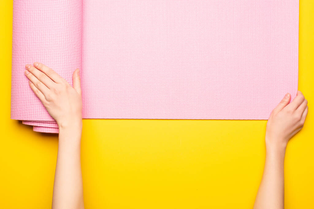 黄色の背景にピンクのフィットネスマットを展開する女性のクロップドビュー - 写真・画像