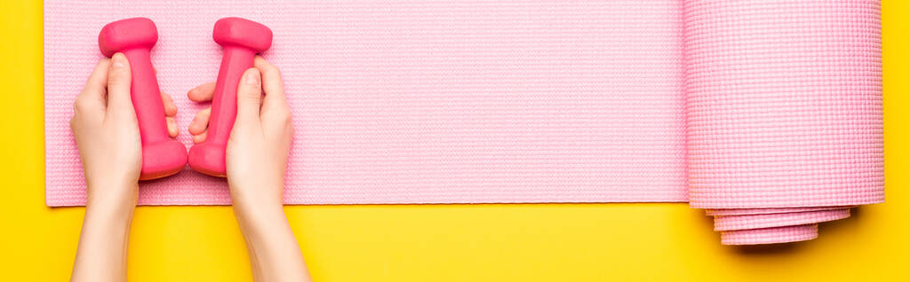 top view női kéz súlyzókkal rózsaszín fitness szőnyeg sárga háttér, panoráma lövés - Fotó, kép
