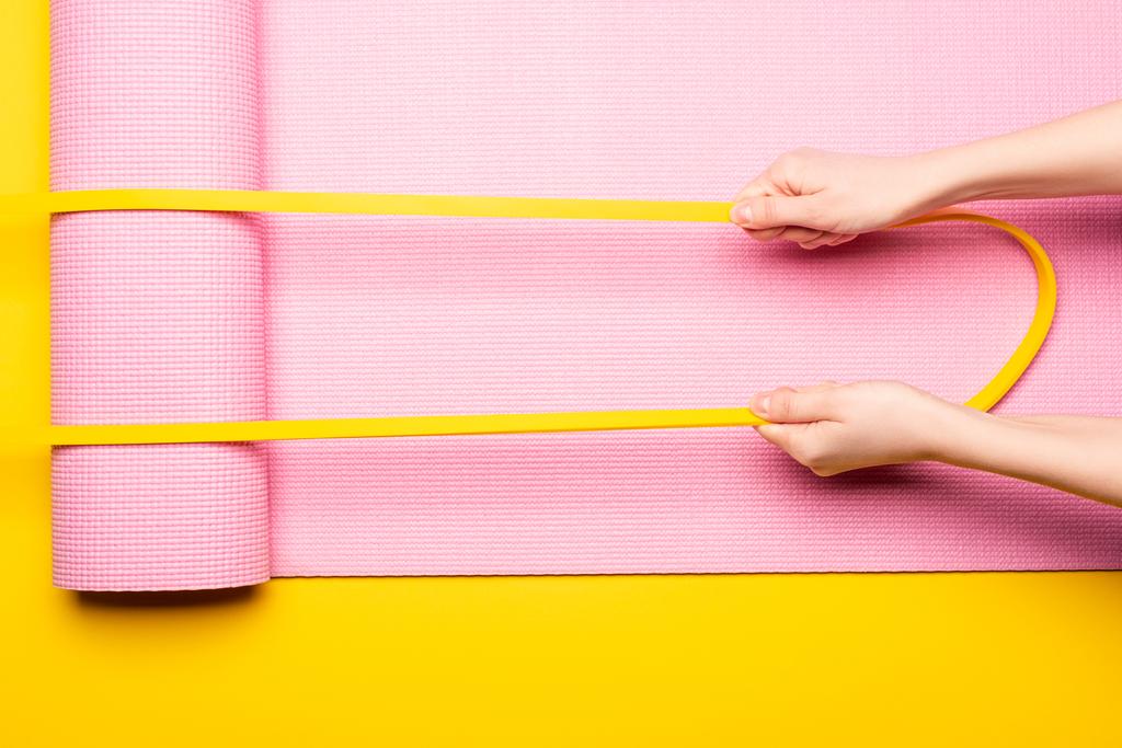 обрезанный вид женщины, держащей упругую ленту на розовом коврике фитнес на желтом фоне - Фото, изображение