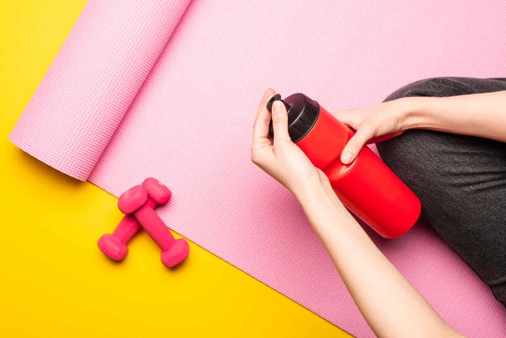 gedeeltelijk uitzicht op vrouw houden sportfles terwijl zitten op roze fitness mat in de buurt halters op gele achtergrond - Foto, afbeelding