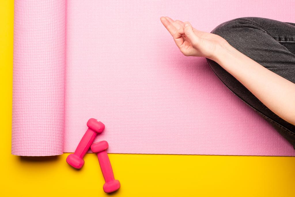 Sarı arka plandaki dambılların yanında pembe fitness minderi üzerinde meditasyon yapan bir kadın görüntüsü. - Fotoğraf, Görsel