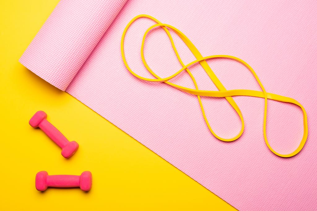 vista superior de la banda de resistencia en la alfombra de fitness rosa cerca de pesas sobre fondo amarillo
 - Foto, Imagen