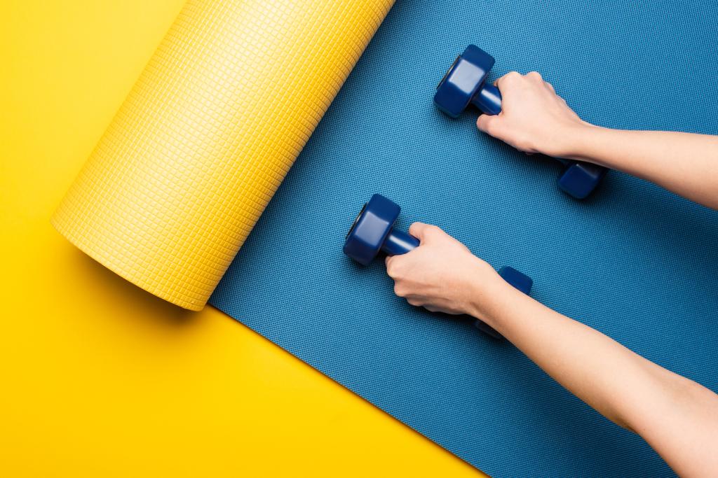 cropped view of woman holding dumbbells on blue fitness mat on yellow background - Photo, Image
