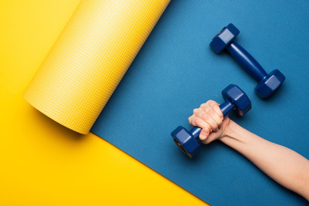 oříznutý pohled na ženu držící činku na modrém fitness podložce na žlutém pozadí - Fotografie, Obrázek