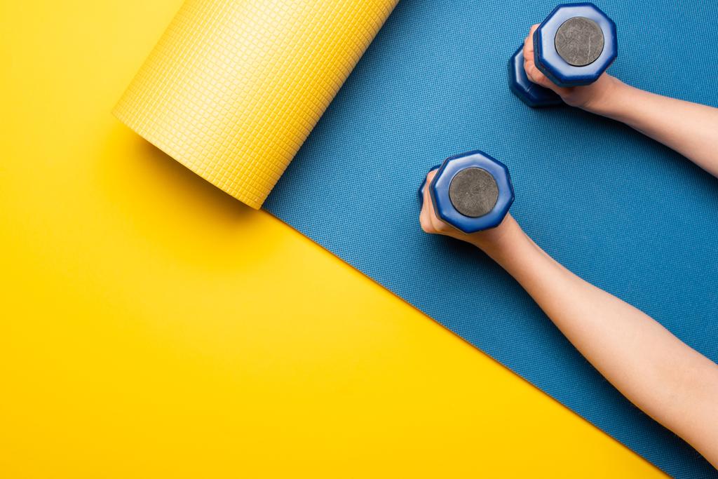 καλλιεργημένη άποψη της γυναίκας που κρατά αλτήρες σε μπλε στρώμα γυμναστικής σε κίτρινο φόντο - Φωτογραφία, εικόνα