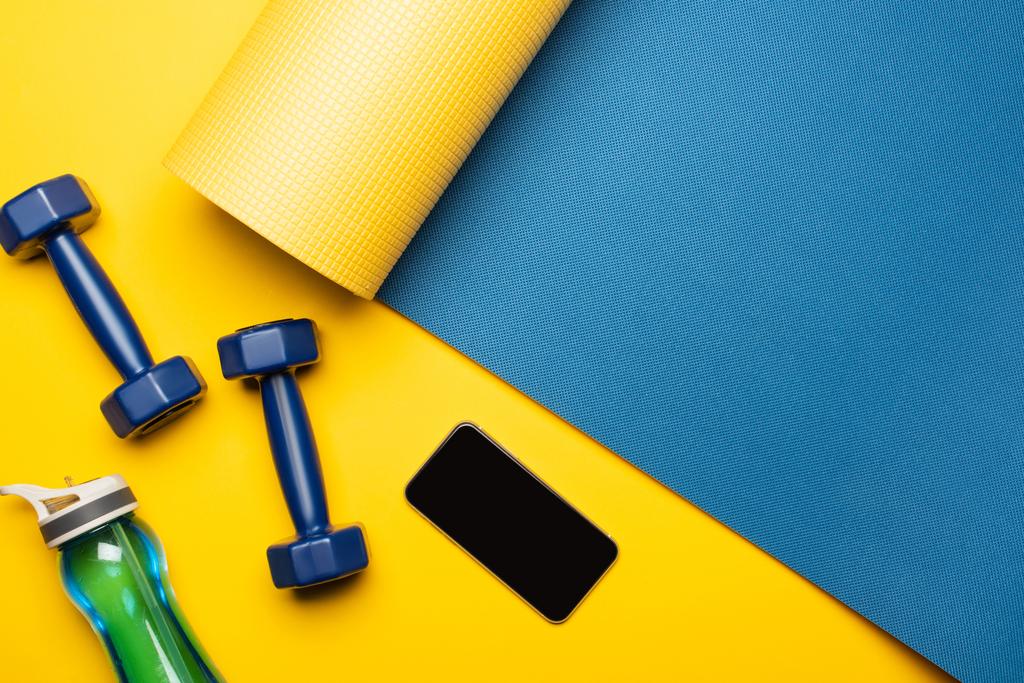 vista dall'alto del tappeto fitness blu con manubri, smartphone e bottiglia sportiva su sfondo giallo - Foto, immagini