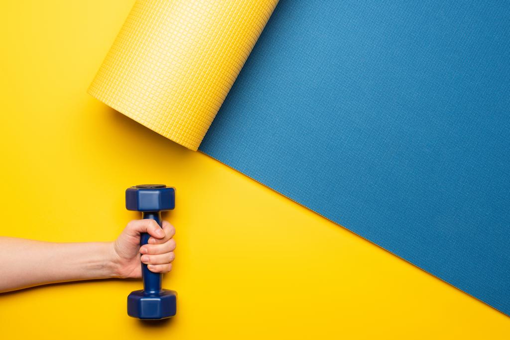 vista ritagliata della donna che tiene i manubri sul tappeto fitness blu su sfondo giallo - Foto, immagini