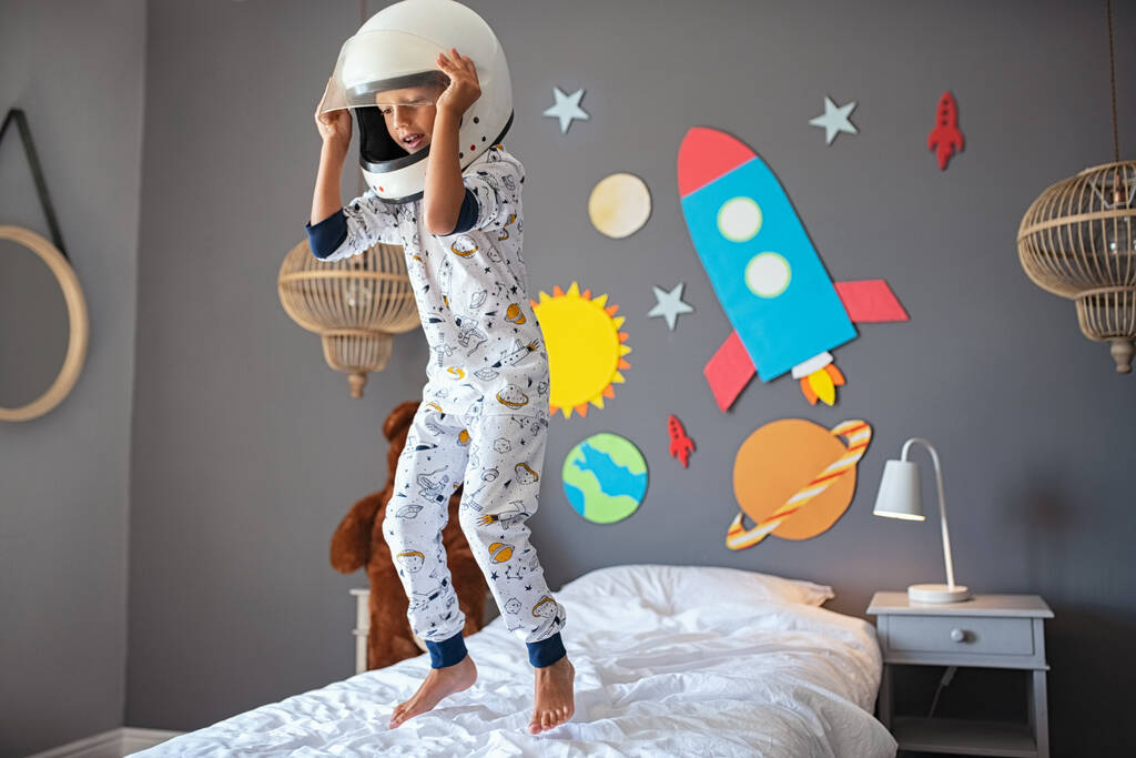 Маленький милый мальчик в костюме космонавта и прыгает на кровати с ракетами и планетами на стене. Ребенок в шлеме астронавта играет и мечтает стать космонавтом. Веселый малыш в космической пижаме и весело проводит время с Солнечной системой и планетами
 - Фото, изображение