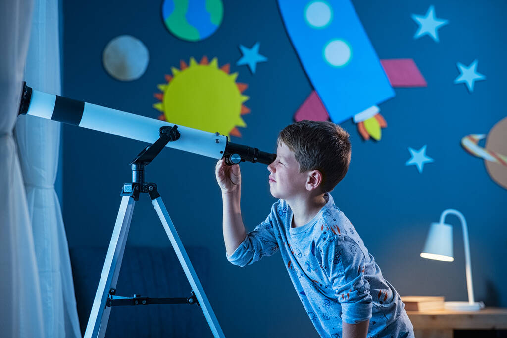 Curieux garçon utilisant le télescope pour explorer la surface de la lune la nuit dans sa chambre. Jeune enfant utilisant le télescope pour voir la galaxie éloignée de la chambre avec mur décoré avec fusée, planètes, étoiles et système solaire en carton. Enfant passionné en pyjama regardant s - Photo, image