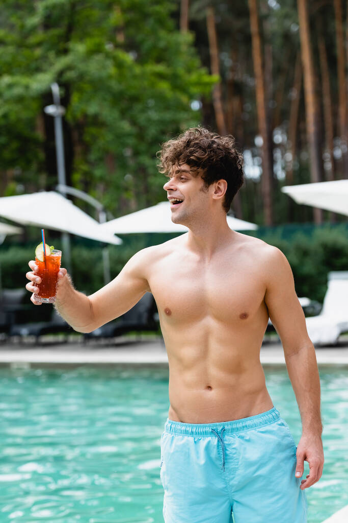 Neşeli ve tişörtsüz bir adam elinde bardakla havuz kenarında kokteyl tutuyor. - Fotoğraf, Görsel