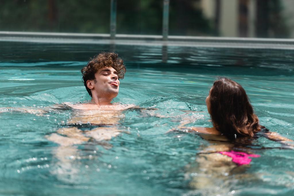 bel homme qui sort la langue tout en nageant avec une femme dans la piscine - Photo, image