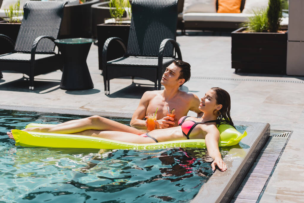 piękna kobieta leżąca na dmuchanym materacu w pobliżu przystojny mężczyzna trzyma koktajl w basenie  - Zdjęcie, obraz