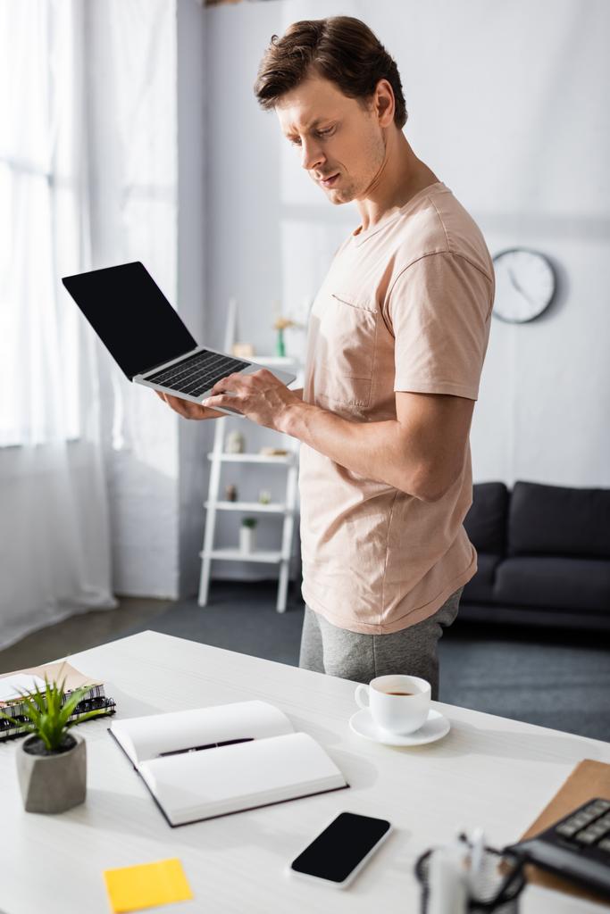 Selective focus of concentrated man using laptop and looking at notebook at home, earning online concept - Photo, Image