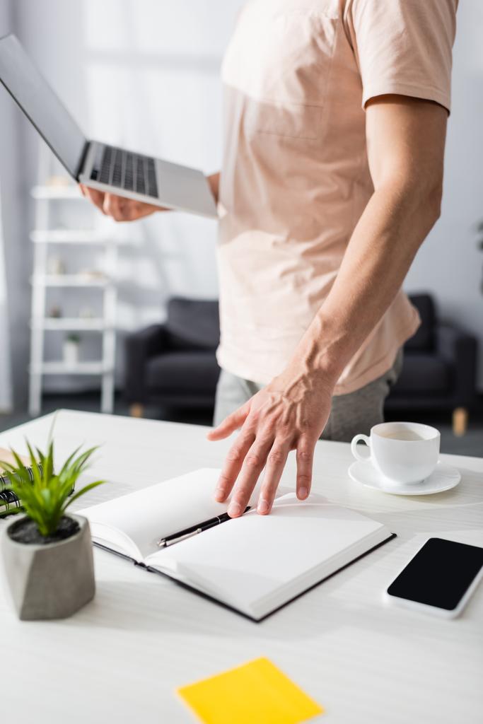 Селективный фокус человека возле ноутбука со смартфоном и заводом на столе дома, зарабатывая онлайн концепцию - Фото, изображение