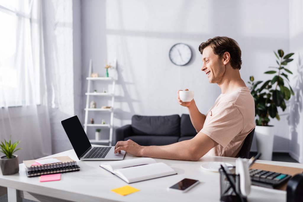 Vista lateral del freelancer sonriente sosteniendo la taza de café y utilizando el ordenador portátil cerca de papelería en la mesa, ganando concepto en línea
 - Foto, Imagen