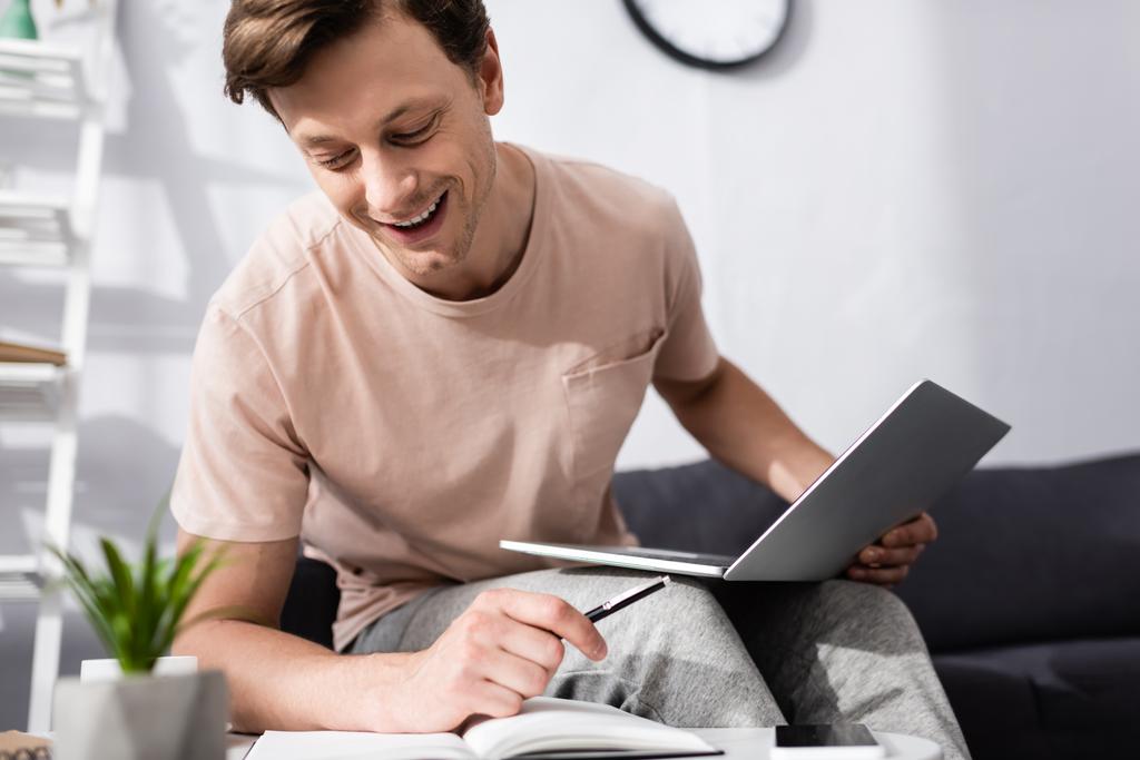 Enfoque selectivo del freelancer sonriente sosteniendo la pluma y el portátil mientras mira el cuaderno en casa, concepto de ganar en línea
 - Foto, imagen