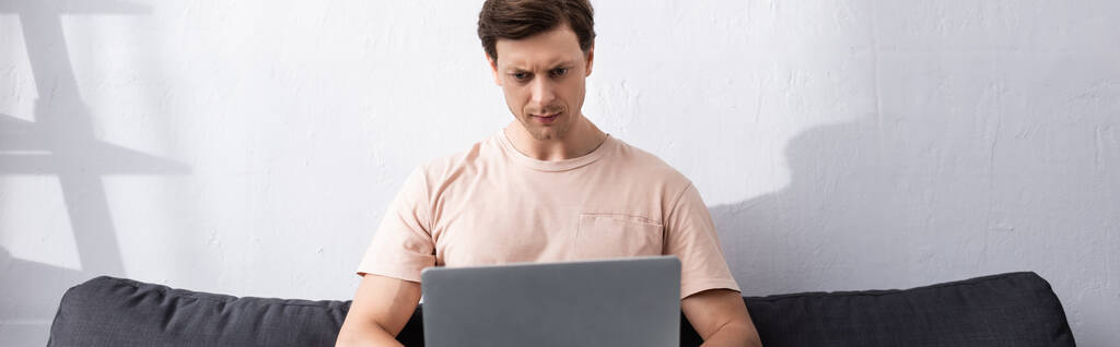 Panoramiczne ujęcie skoncentrowanego mężczyzny korzystającego z laptopa na kanapie w domu, zarabiającego koncepcję online - Zdjęcie, obraz