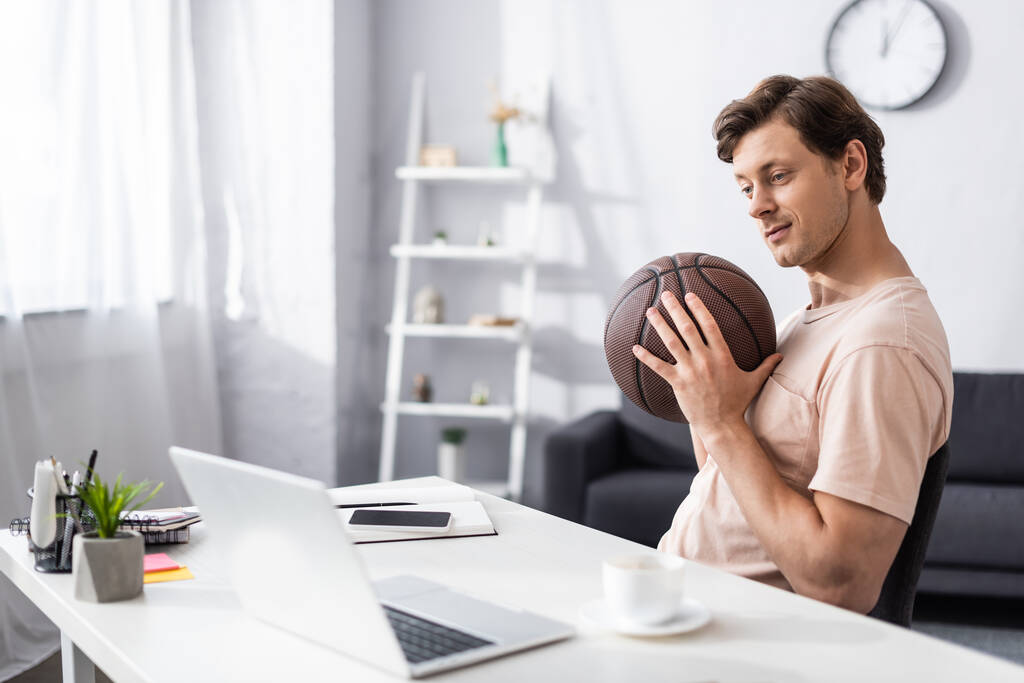 Szelektív fókusz jóképű férfi gazdaság kosárlabda közelében laptop és írószer az asztalon otthon, koncepciója kereső online - Fotó, kép