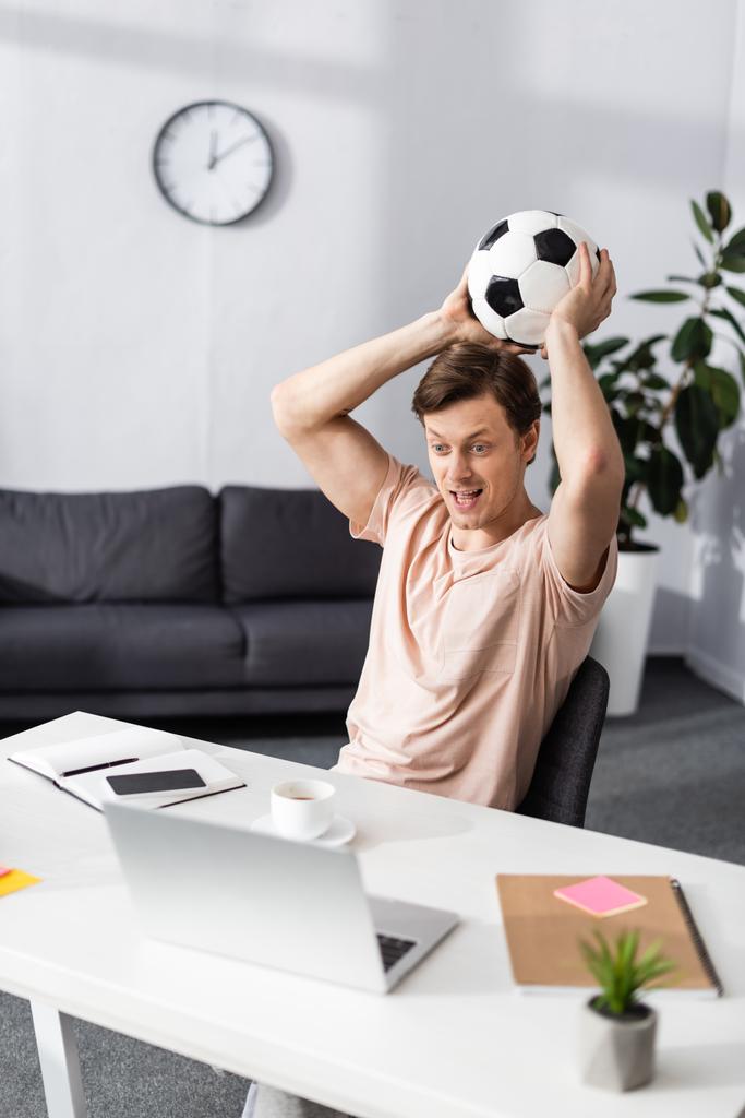 Selektivní zaměření pozitivní muž drží fotbal a při pohledu na notebook na stole doma, koncept vydělávat on-line - Fotografie, Obrázek