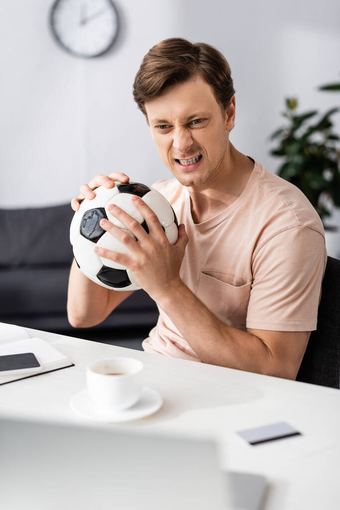 Foco seletivo de homem com raiva segurando futebol perto de gadgets e cartão de crédito na mesa, conceito de ganhar on-line
 - Foto, Imagem