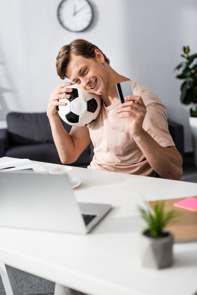 Selektiver Fokus eines lächelnden Mannes, der Kreditkarte und Fußball in der Nähe des Laptops auf dem Tisch hält und Online-Konzept verdient - Foto, Bild