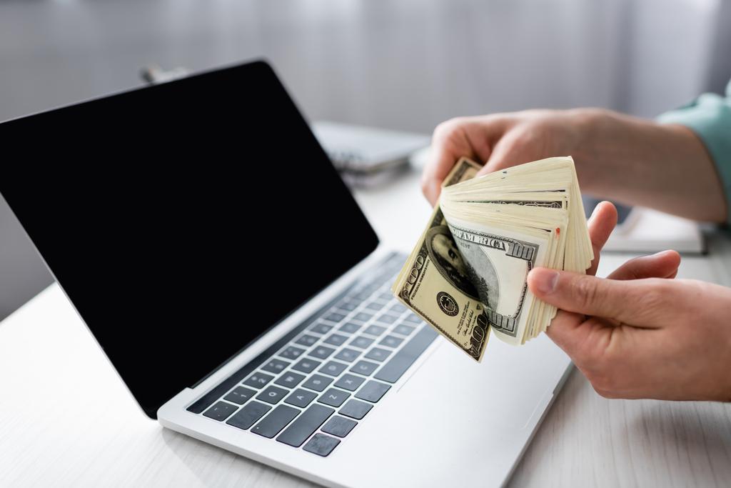 Rajatut näkymät mies pitää dollarin seteleitä lähellä kannettavaa tietokonetta tyhjä näyttö pöydällä, ansaita verkossa käsite - Valokuva, kuva