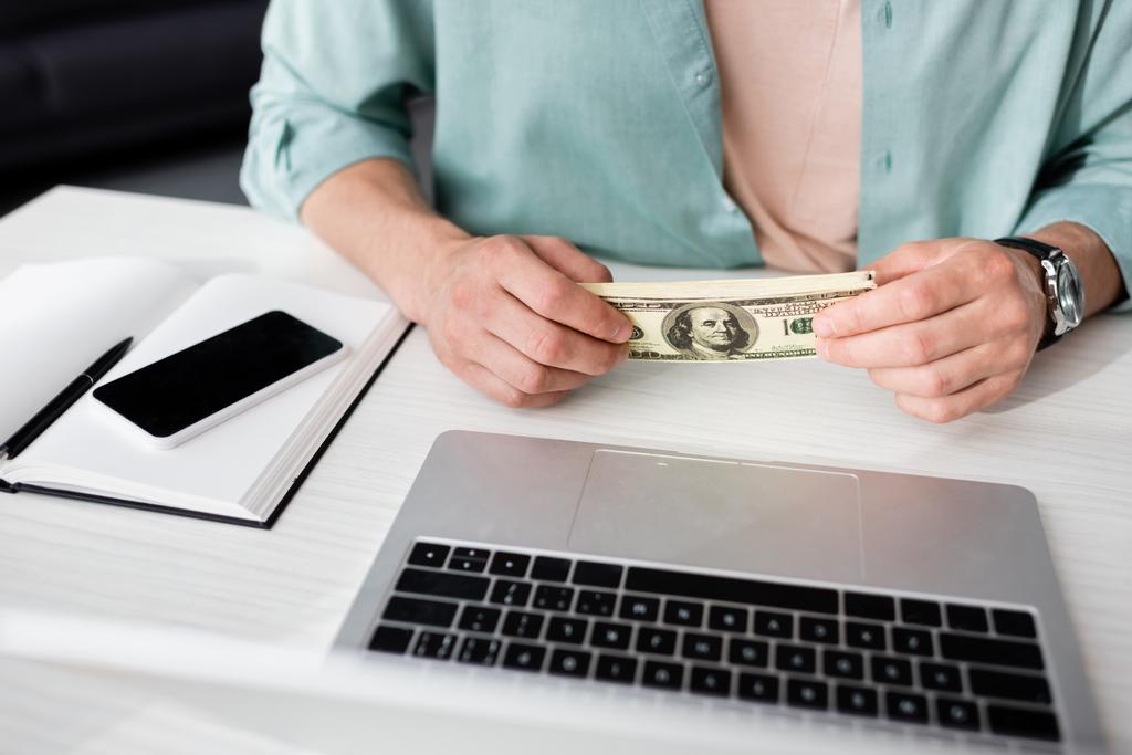 Vista ritagliata di uomo in possesso di dollari vicino laptop e smartphone con schermo bianco sul tavolo, concetto di guadagnare online - Foto, immagini