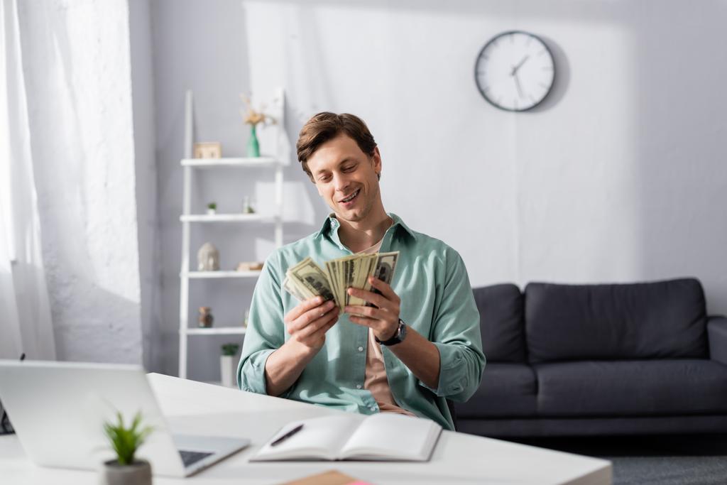 Selective focus of smiling man counting money near laptop and notebook on table in living room, concept of earning online - Photo, Image