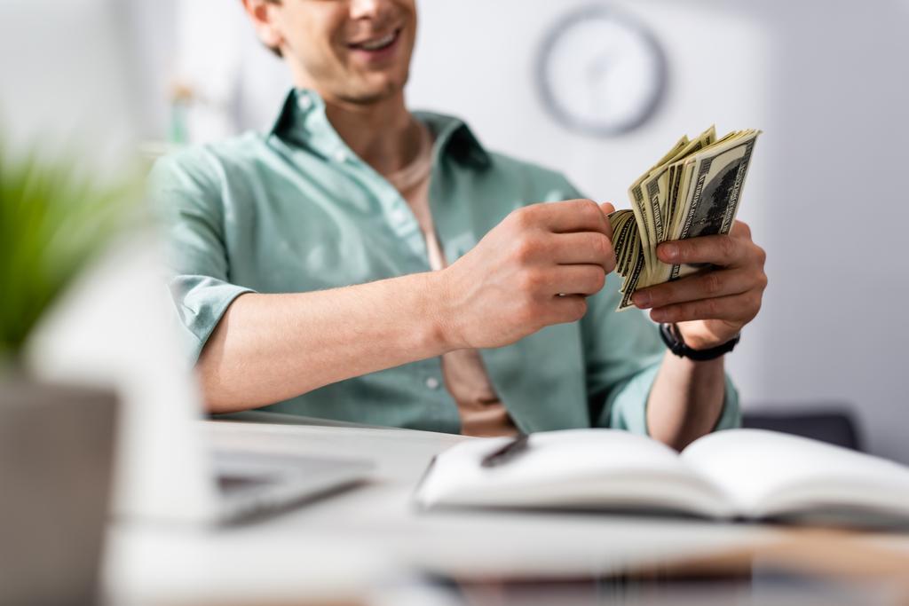 Περικοπή άποψη του χαμογελαστού freelancer καταμέτρηση δολάρια κοντά φορητό υπολογιστή και σημειωματάριο στο τραπέζι, κερδίζοντας online έννοια - Φωτογραφία, εικόνα