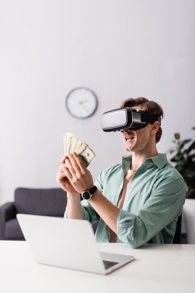 Concentration sélective de l'homme souriant dans le casque vr tenant de l'argent près de l'ordinateur portable sur la table, concept de gagner en ligne - Photo, image