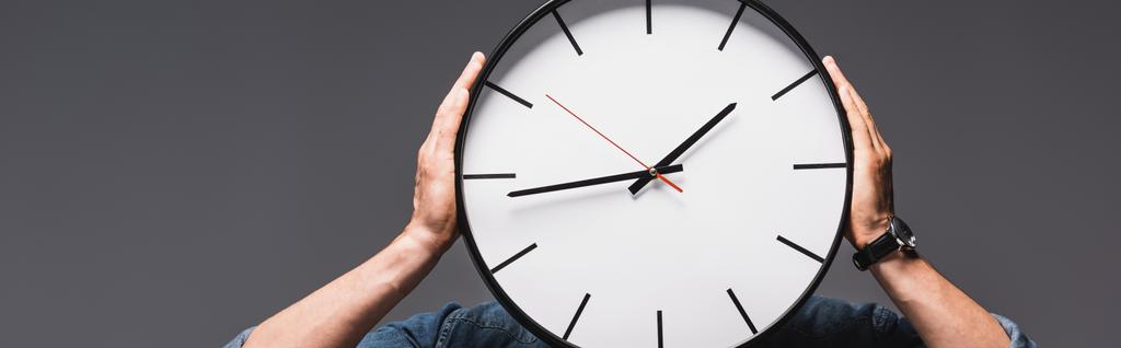 灰色で隔離された顔の近くに時計を保持する男のパノラマ作物、時間管理の概念  - 写真・画像