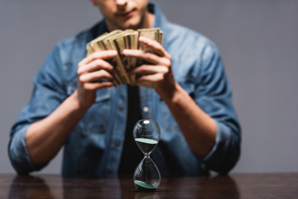 Selective focus of hourglass on table and man holding money isolated on grey, concept of time management  - Photo, Image