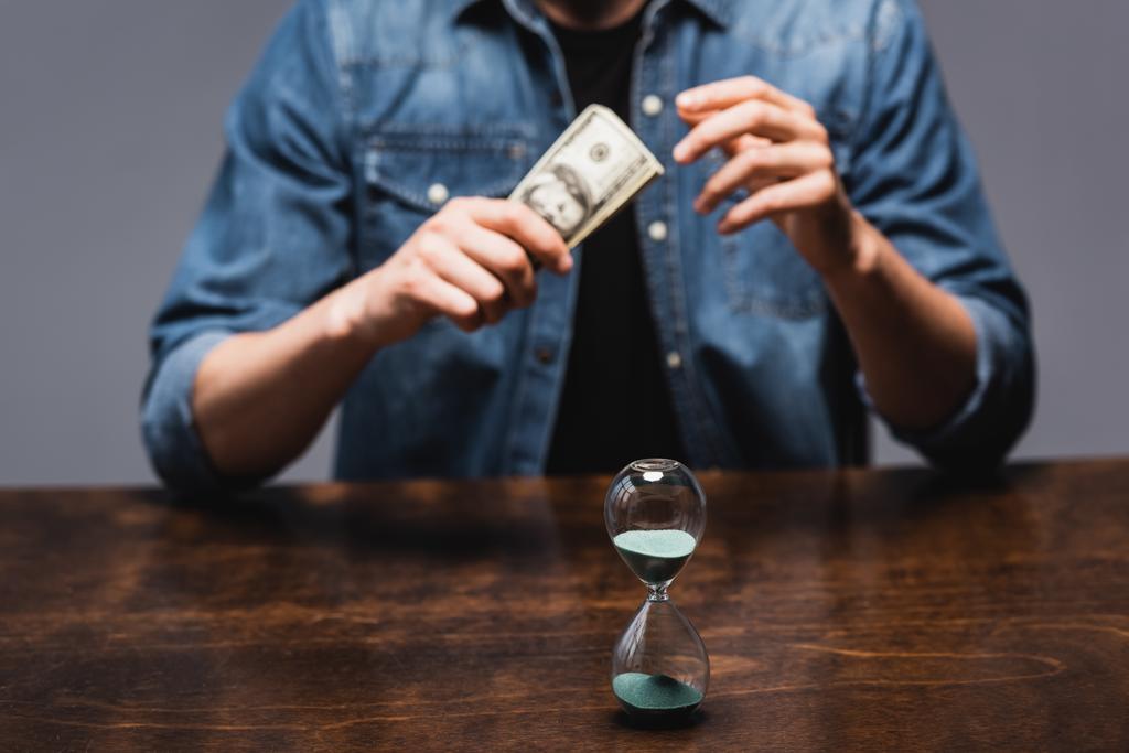 Foco seletivo de ampulheta perto de homem segurando dólares enquanto sentado à mesa isolado em cinza, conceito de gerenciamento de tempo
  - Foto, Imagem
