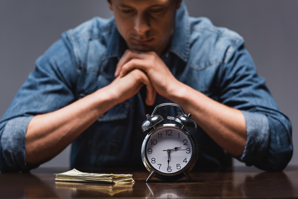 Foco seletivo de dinheiro e despertador na mesa perto de homem pensativo isolado em cinza, conceito de gerenciamento de tempo
  - Foto, Imagem