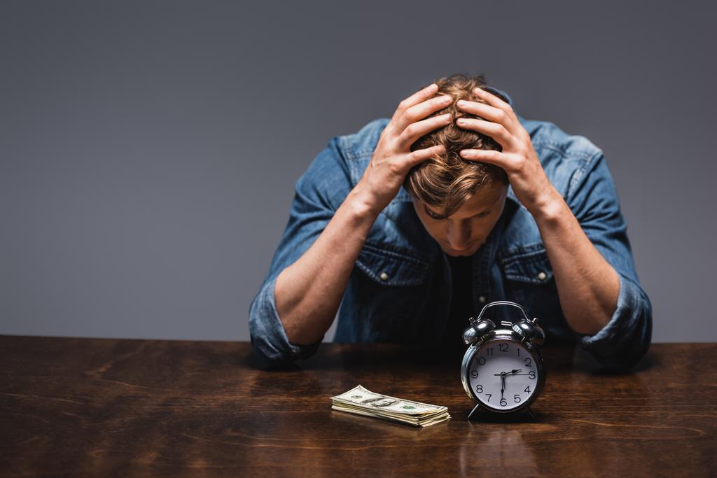 Недовольный человек смотрит на деньги и будильник на столе изолированы на серый, концепция управления временем  - Фото, изображение