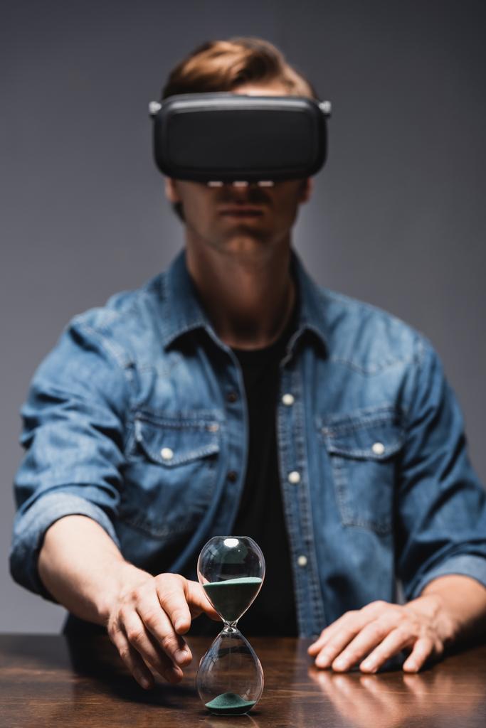 Επιλεκτική εστίαση του ανθρώπου στην εικονική πραγματικότητα ακουστικά τραβώντας το χέρι σε κλεψύδρα στο τραπέζι σε γκρι φόντο, έννοια της διαχείρισης του χρόνου  - Φωτογραφία, εικόνα