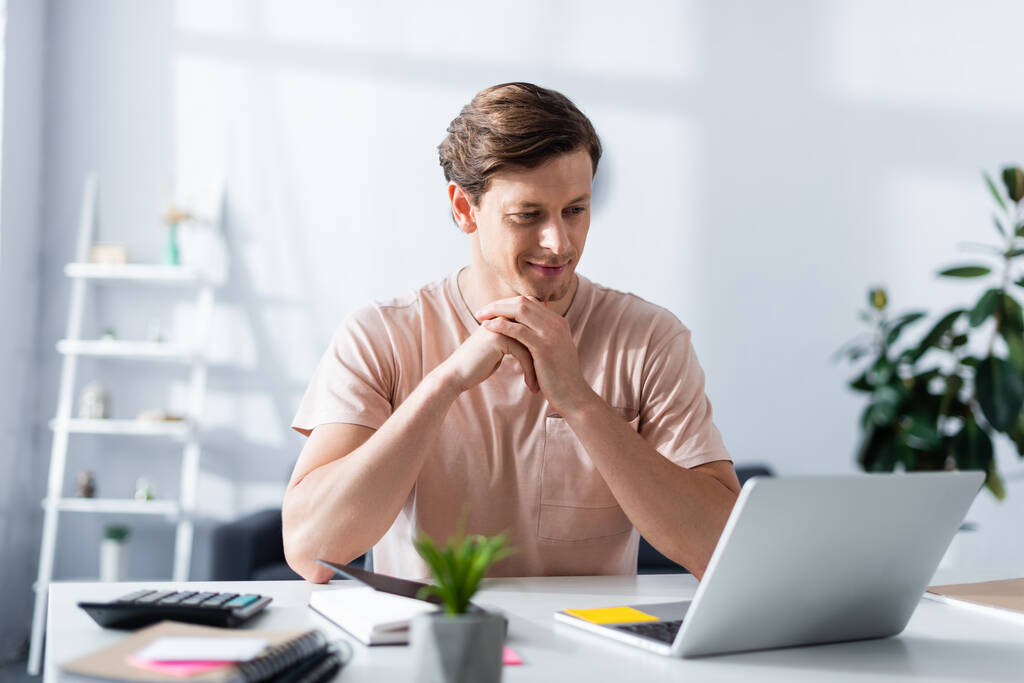 Вибірковий фокус усміхненого чоловіка, який дивиться на ноутбук біля канцтоварів на столі вдома, заробляє онлайн-концепцію
 - Фото, зображення
