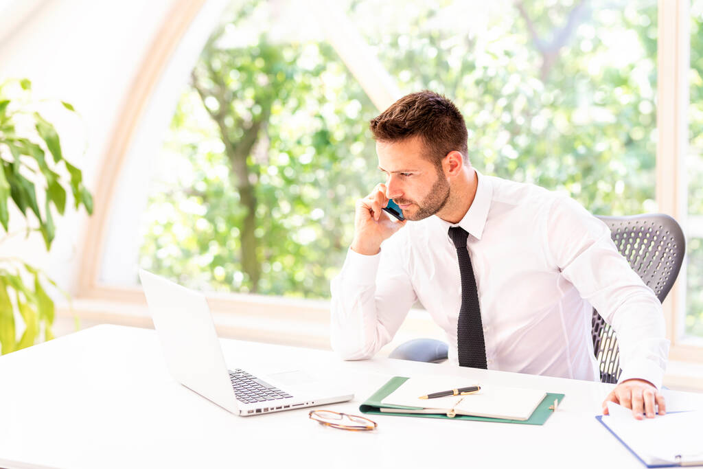 Портрет красивого бізнесмена, який розмовляє з кимось на мобільному телефоні і використовує ноутбук, сидячи за столом в офісі
.  - Фото, зображення