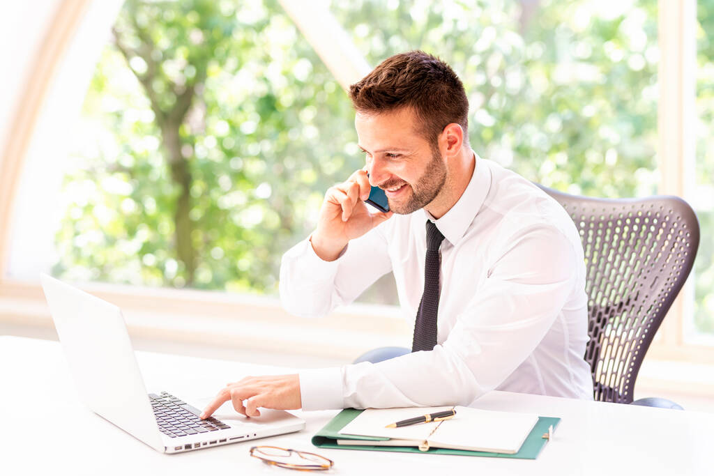 Portret przystojnego biznesmena rozmawiającego z kimś przez telefon komórkowy i korzystającego z laptopa siedząc przy biurku w biurze.  - Zdjęcie, obraz