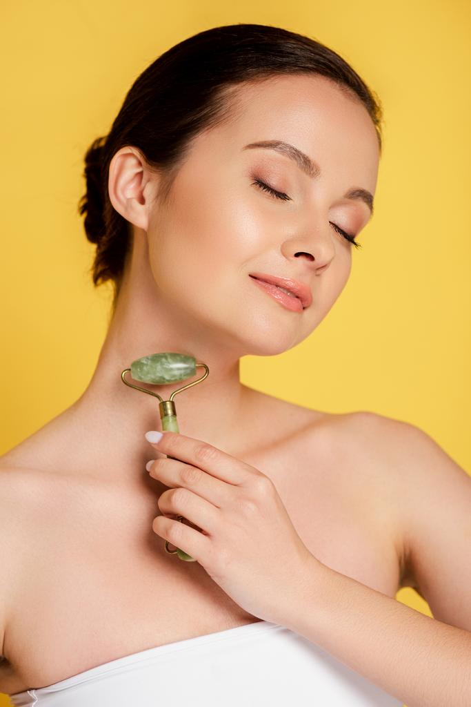 beautiful woman with closed eyes using jade roller on face isolated on yellow - Photo, Image