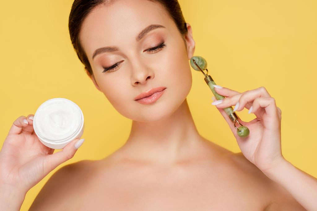 naked beautiful woman holding cosmetic cream and jade roller isolated on yellow - Photo, Image
