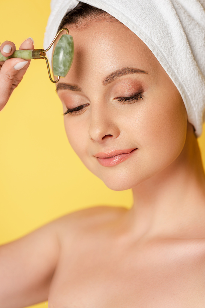 beautiful naked woman with towel on head using jade roller isolated on yellow - Photo, Image