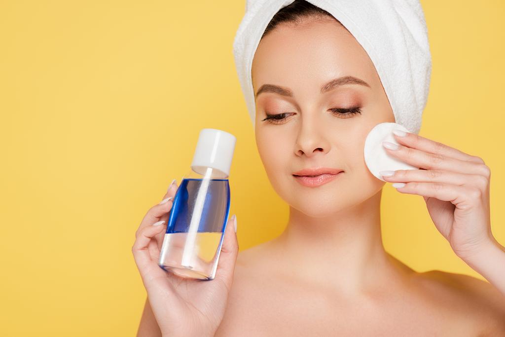 beautiful naked woman with towel on head, micellar water and cotton pad isolated on yellow - Photo, Image