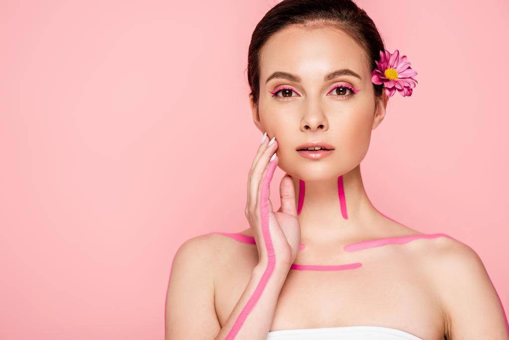 schöne Frau mit rosa Linien auf dem Körper und Blume im Haar berühren Gesicht isoliert auf rosa - Foto, Bild