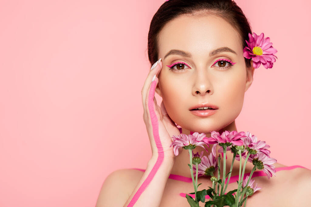 γυμνή όμορφη γυναίκα με ροζ γραμμές στο σώμα και το λουλούδι στα μαλλιά κρατώντας μπουκέτο και αγγίζοντας το πρόσωπο απομονώνονται σε ροζ - Φωτογραφία, εικόνα