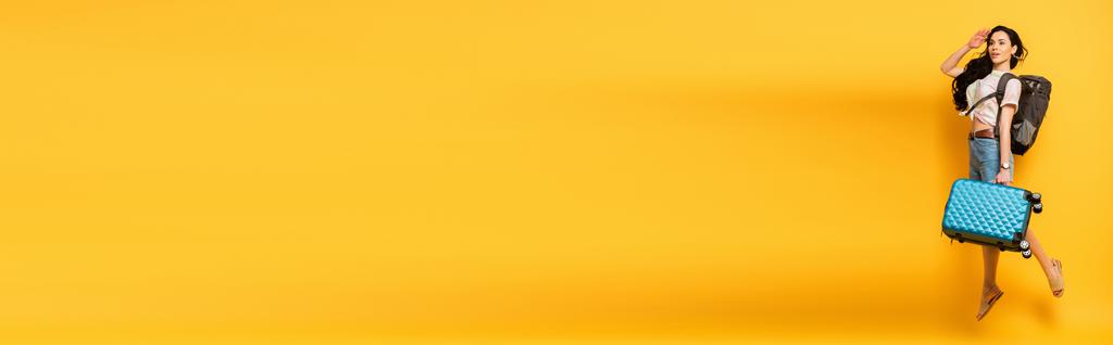 брюнетка женщина прыгает с рюкзаком и чемоданом и смотрит в сторону на желтом фоне, панорамный снимок - Фото, изображение