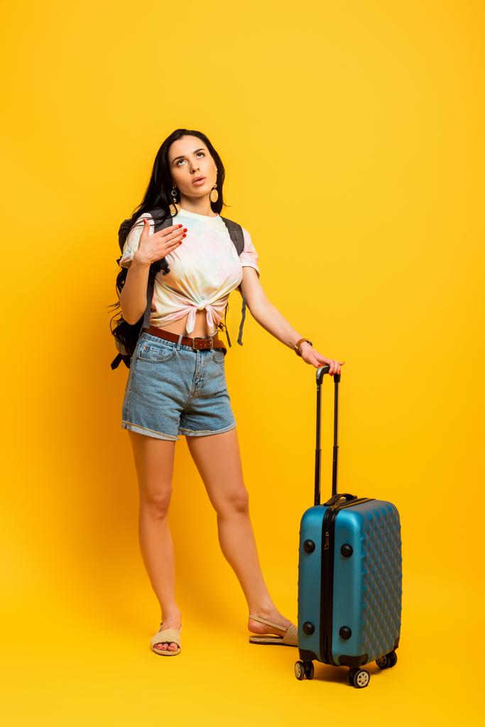 brunette vrouw met rugzak en koffer lijden aan warmte op gele achtergrond - Foto, afbeelding