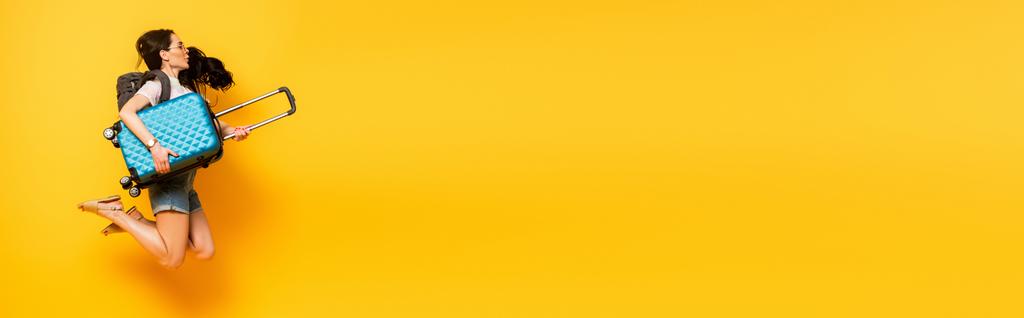 боковой вид брюнетки, прыгающей с рюкзаком и чемоданом на желтом фоне, панорамный снимок - Фото, изображение