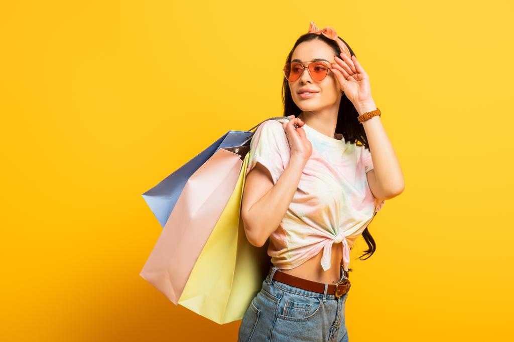 lächelnd stilvolle Sommer Brünette Mädchen mit Einkaufstaschen Blick weg auf gelbem Hintergrund - Foto, Bild