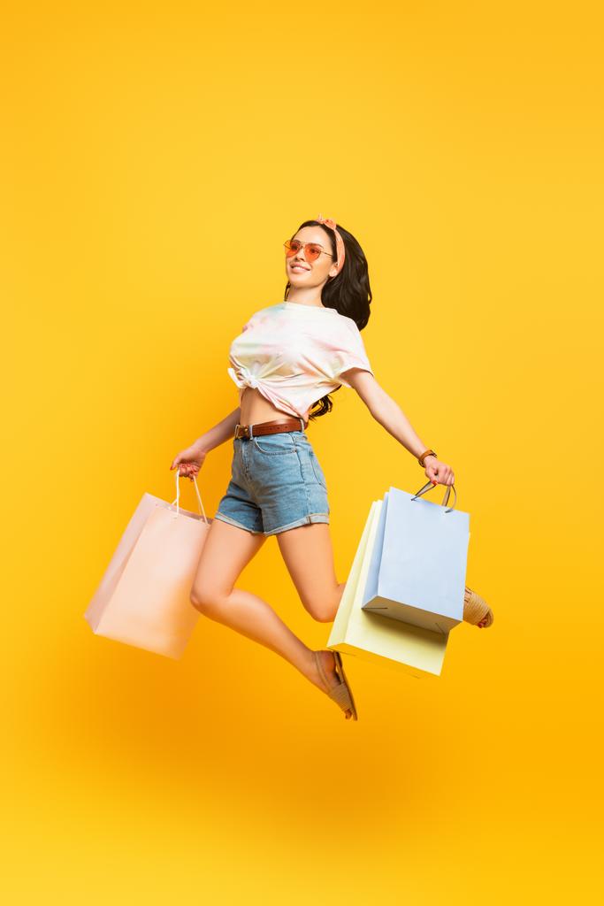 Вид в полный рост улыбающейся стильной летней брюнетки, прыгающей с сумками для покупок на желтом фоне - Фото, изображение